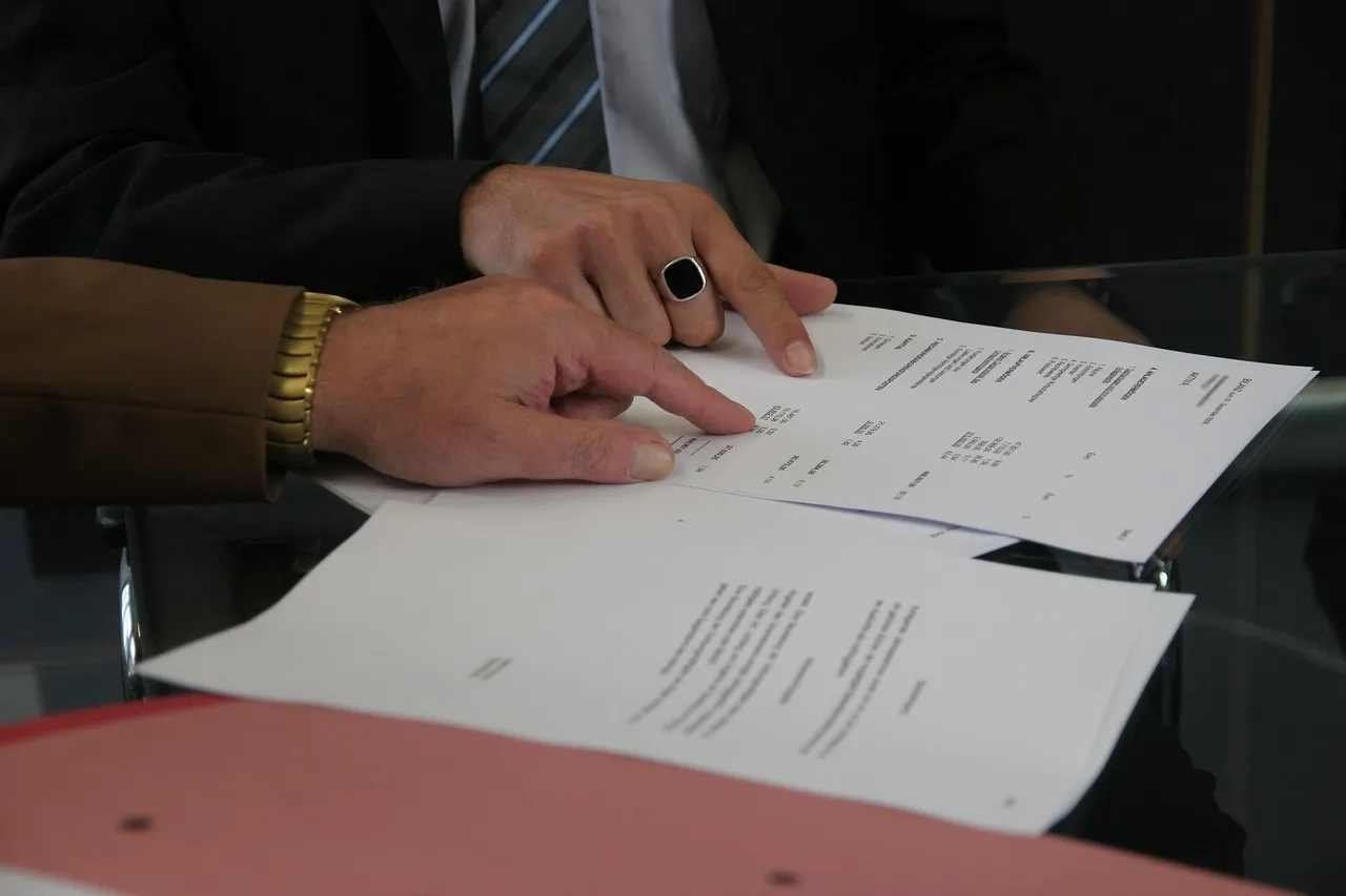 The image shows two hands examining a stack of white documents on a glossy table, with a man in a suit partially visible in the background. The scene appears to be an office or meeting room, suggested by the reflective surface beneath the p — jail house notary in Arlington, TX